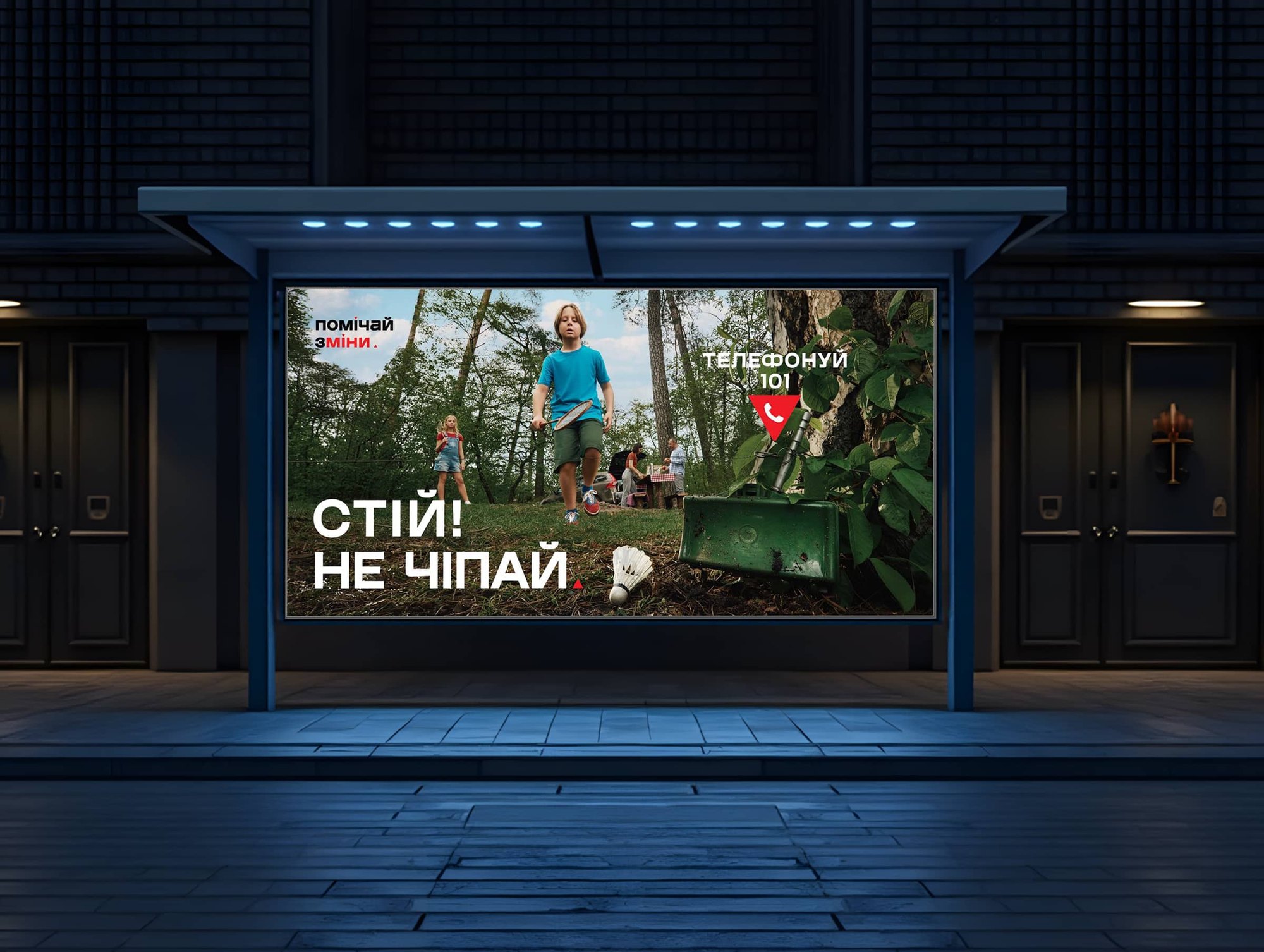 Campaign billboard at a night bus stop: children playing badminton near a hidden mine — “Stop! Don’t touch!”. Call 101